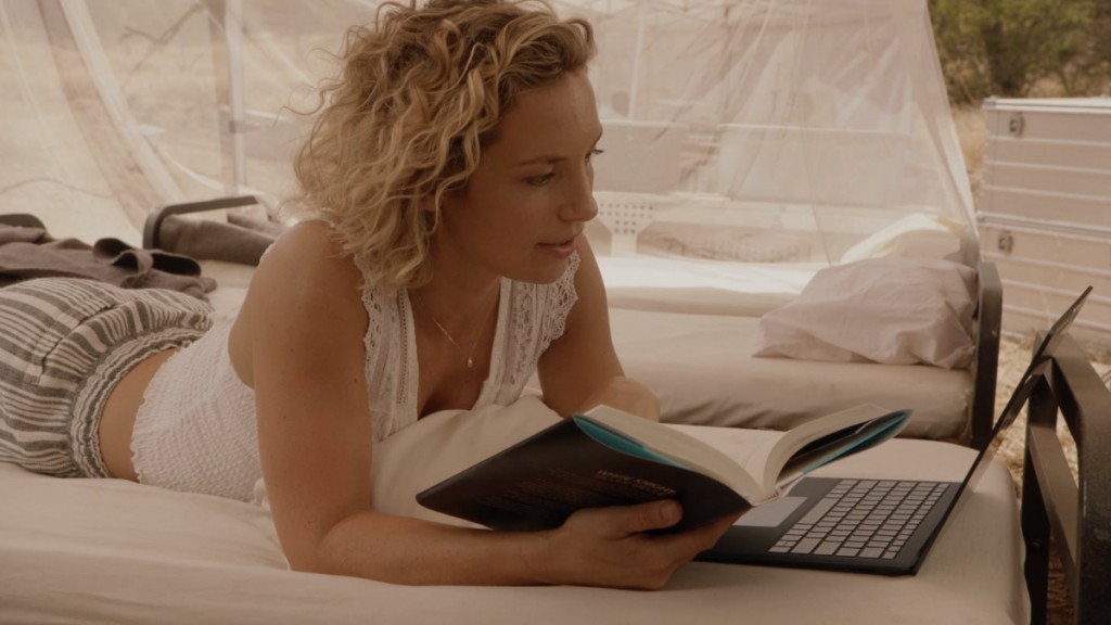 Tout en tenant un livre à la main, Higgins (Perdita Weeks) regarde quelque chose sur son ordinateur.